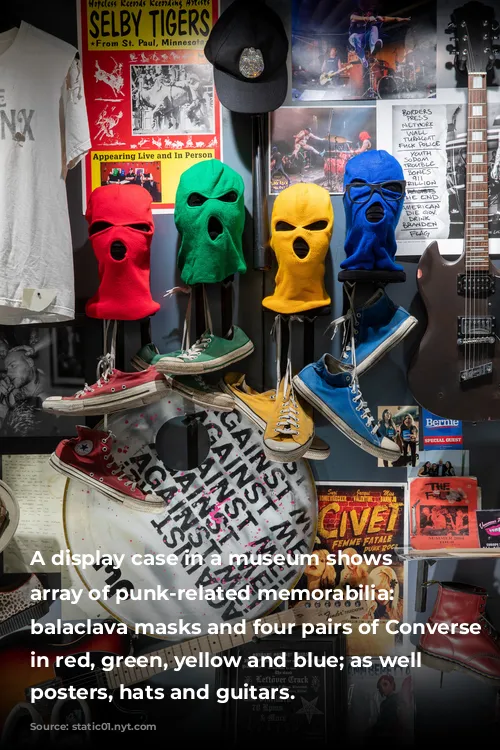 A display case in a museum shows an array of punk-related memorabilia: Four balaclava masks and four pairs of Converse sneakers in red, green, yellow and blue; as well as posters, hats and guitars.