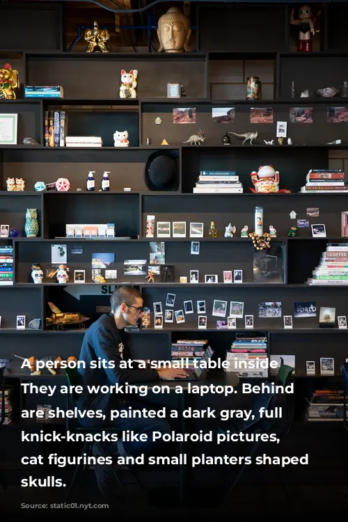 A person sits at a small table inside cafe. They are working on a laptop. Behind them are shelves, painted a dark gray, full of knick-knacks like Polaroid pictures, waving cat figurines and small planters shaped like skulls. 