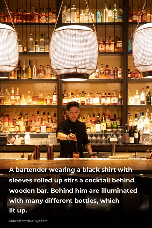 A bartender wearing a black shirt with the sleeves rolled up stirs a cocktail behind a wooden bar. Behind him are illuminated shelves with many different bottles, which are lit up. 