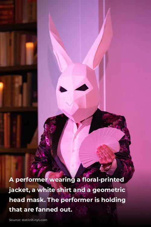 A performer wearing a floral-printed lounge jacket, a white shirt and a geometric rabbit head mask. The performer is holding cards that are fanned out. 