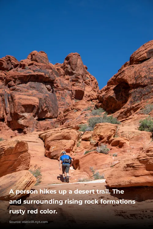 A person hikes up a desert trail. The trail and surrounding rising rock formations are rusty red color. 