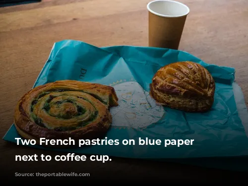 Two French pastries on blue paper bag next to coffee cup.