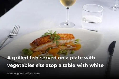 A grilled fish served on a plate with sauteed vegetables sits atop a table with white tablecloth.