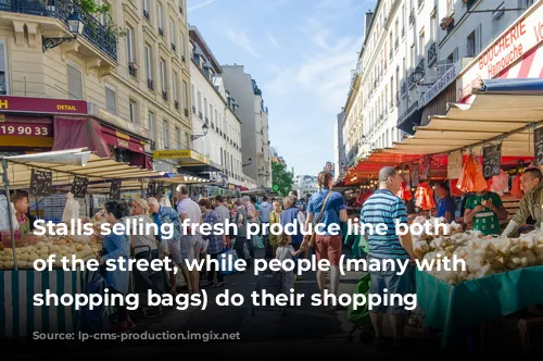 Stalls selling fresh produce line both sides of the street, while people (many with wheelie shopping bags) do their shopping