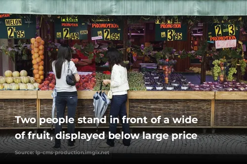 Two people stand in front of a wide range of fruit, displayed below large price signs