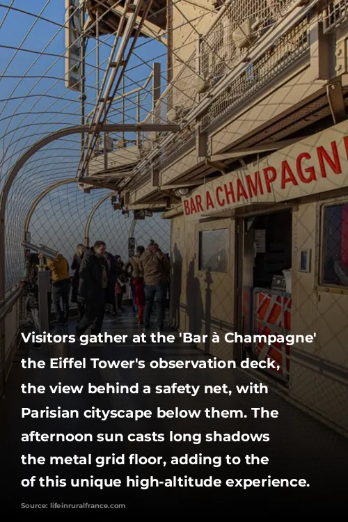 Visitors gather at the 'Bar à Champagne' on the Eiffel Tower's observation deck, enjoying the view behind a safety net, with the Parisian cityscape below them. The late afternoon sun casts long shadows across the metal grid floor, adding to the ambiance of this unique high-altitude experience.