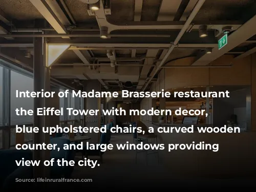 Interior of Madame Brasserie restaurant at the Eiffel Tower with modern decor, featuring blue upholstered chairs, a curved wooden bar counter, and large windows providing a view of the city.