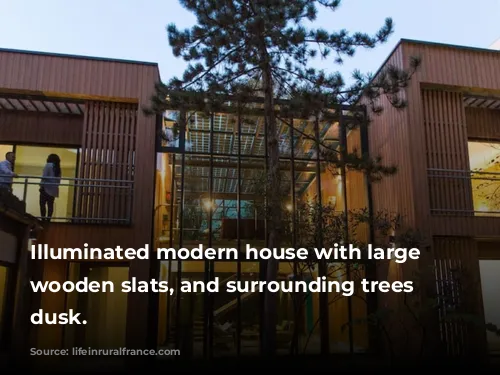 Illuminated modern house with large windows, wooden slats, and surrounding trees at dusk.