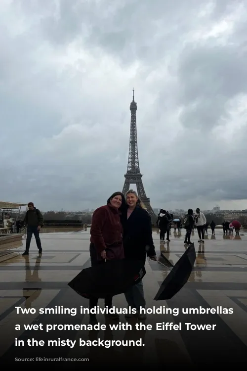 Two smiling individuals holding umbrellas on a wet promenade with the Eiffel Tower rising in the misty background.